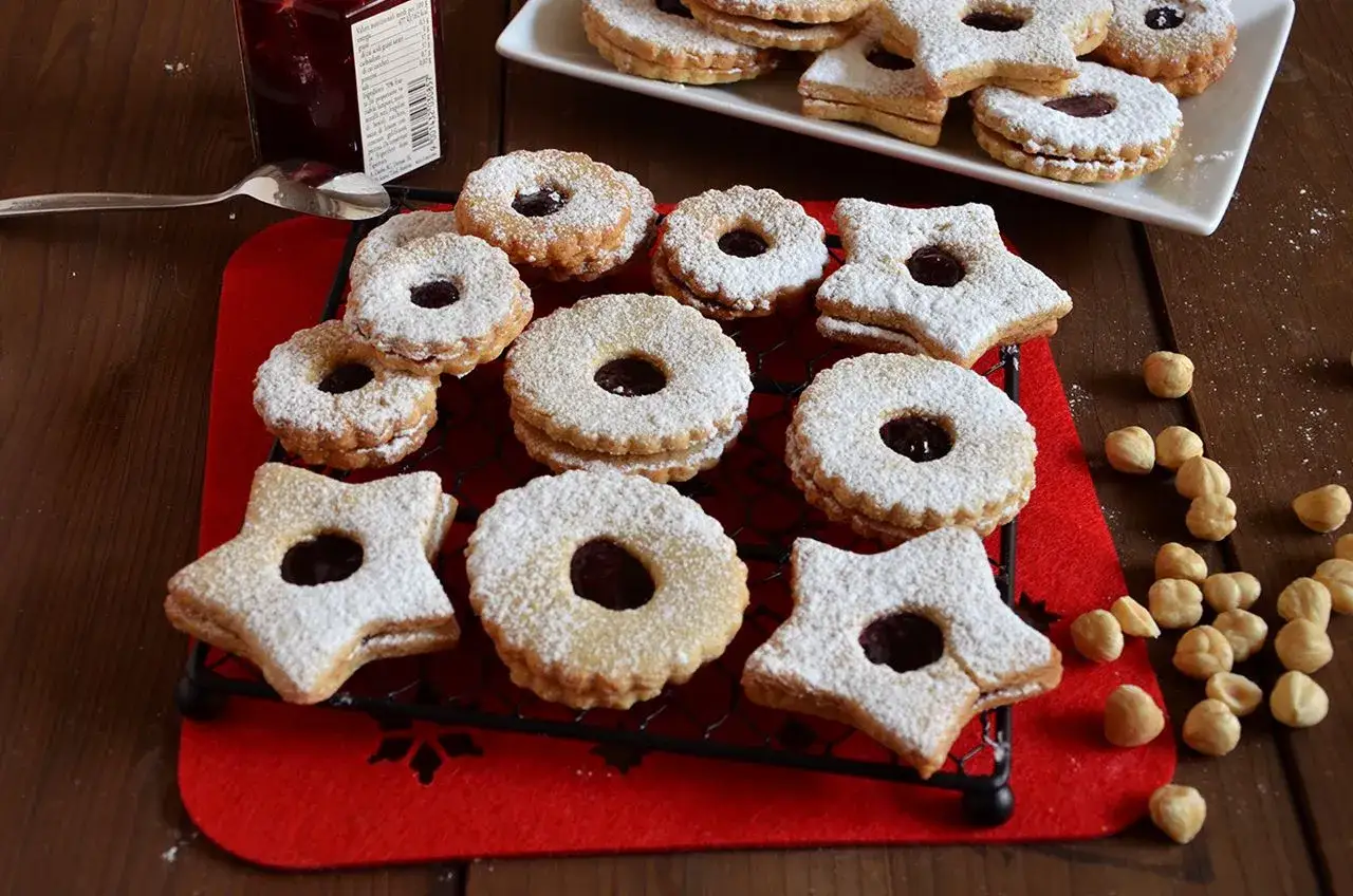 Biscotti Linzer cosparsi di zucchero a velo, ripieni di marmellata, su una griglia rossa. Alcune nocciole sparse.