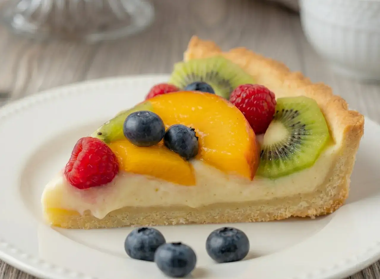 Fetta di crostata con pasta frolla per crostata di frutta, ricca di crema e frutta fresca come kiwi, pesche, lamponi e mirtilli.