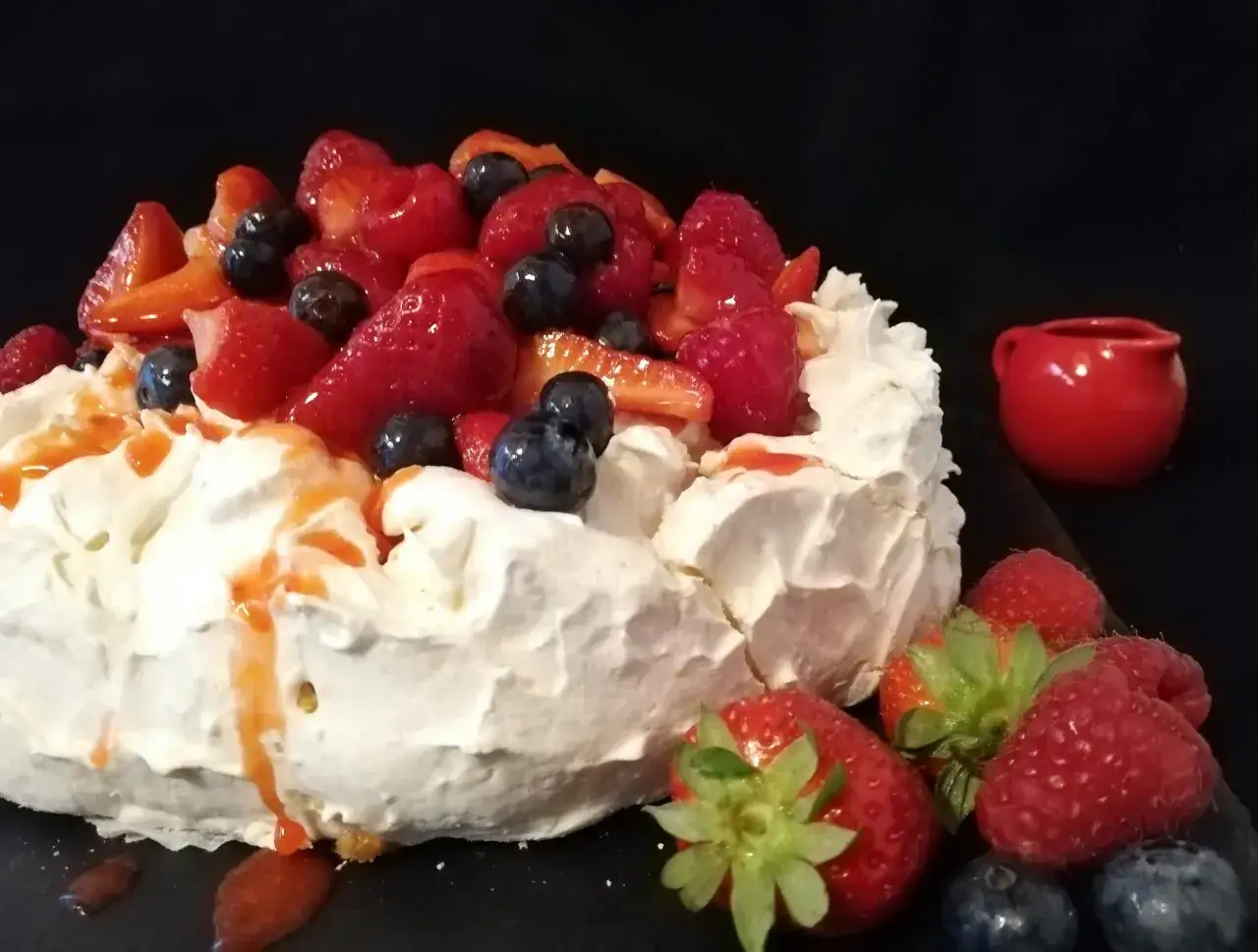 Pavlova ricetta: meringa croccante, panna montata e frutti di bosco freschi, con un tocco di salsa rossa.