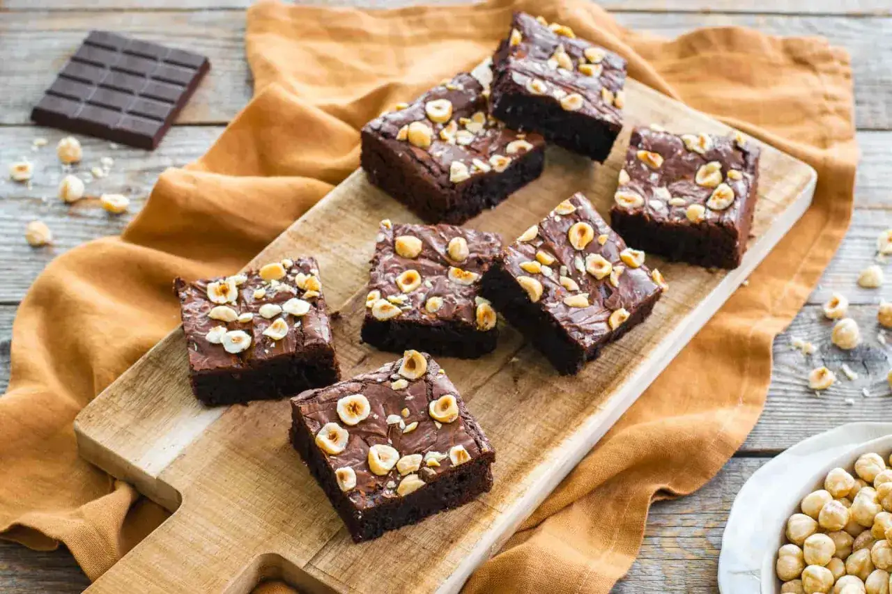 Deliziosi brownie senza glutine con nocciole tostate, disposti su un tagliere di legno, accanto a una tavoletta di cioccolato e una ciotola di nocciole.