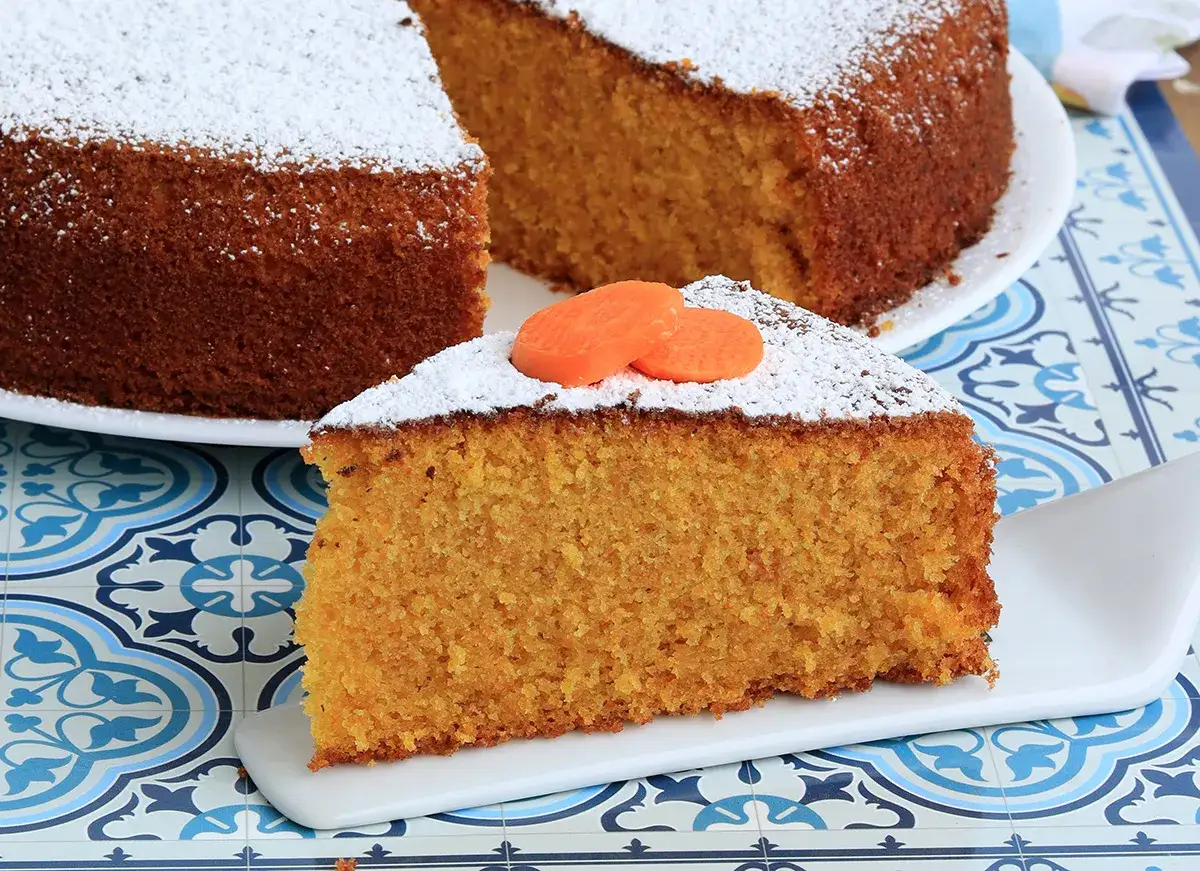 Una fetta di torta di carote senza glutine, soffice e profumata, cosparsa di zucchero a velo e decorata con fettine di carota.