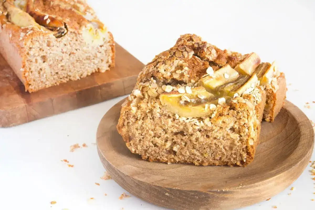 Fetta di banana bread vegano, guarnita con fiocchi d'avena e fettine di banana. Un altro pezzo di torta è sullo sfondo.