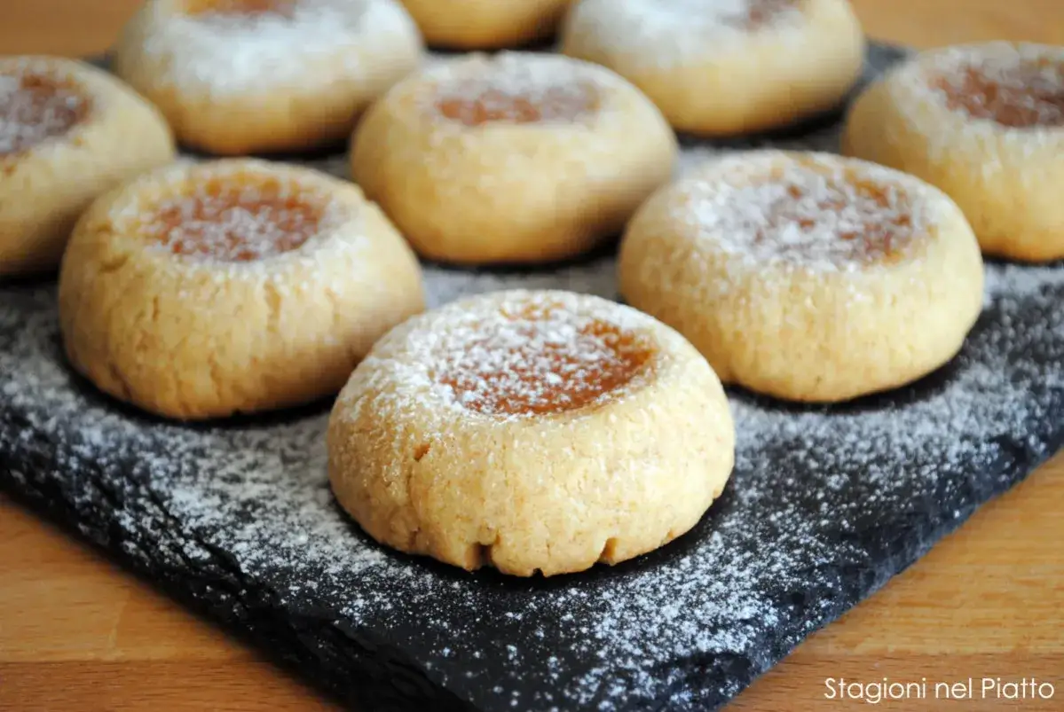 Biscotti integrali con marmellata, spolverati di zucchero a velo, su una lavagna nera.