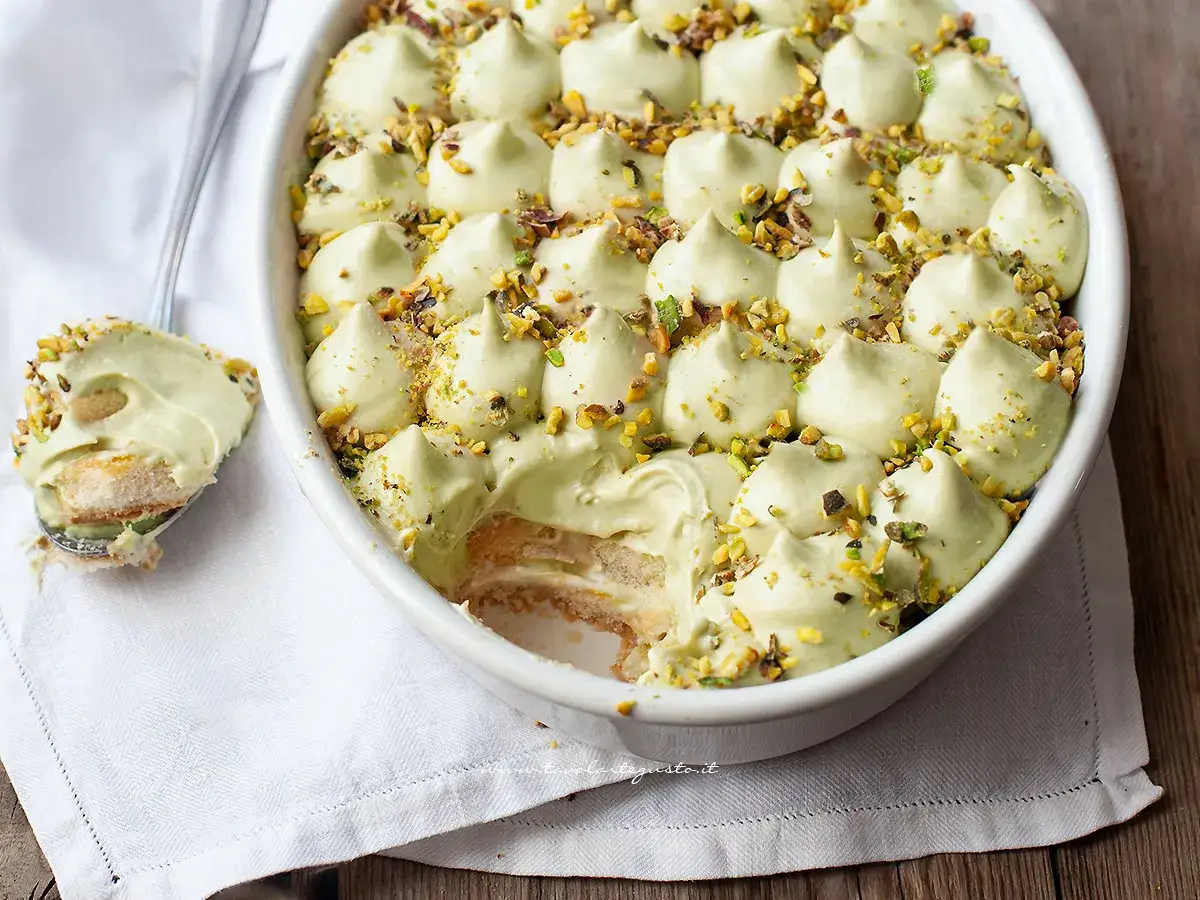 Un cremoso tiramisù al pistacchio, guarnito con granella di pistacchi, in una teglia bianca. Un cucchiaio ne preleva una porzione invitante.