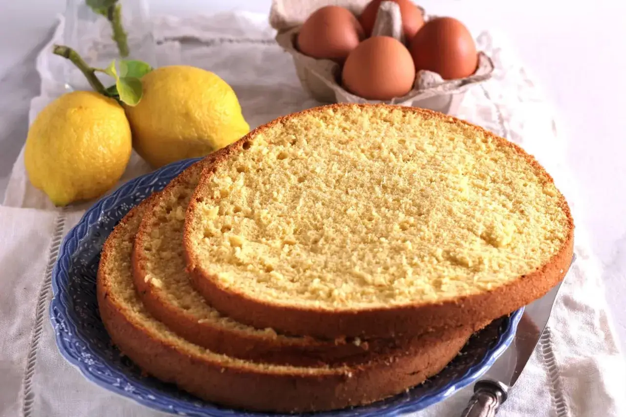 Fette di pan di spagna soffice su un piatto blu, con limoni e uova sullo sfondo.