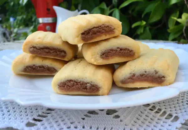 Biscotti nascondini impilati su un piatto bianco, con un ripieno scuro visibile. Sullo sfondo, una moka rossa e foglie verdi.