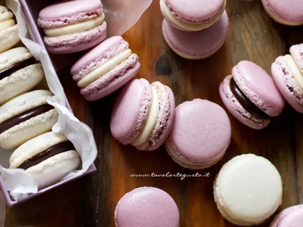 Deliziosi macaron dolci rosa e bianchi, alcuni con ripieno al cioccolato, sparsi su un tagliere di legno.