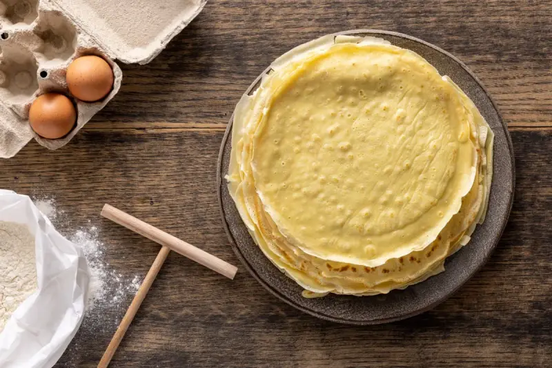 Pila di crepes dorate, pronte per essere farcite. Accanto, uova fresche e farina, ingredienti perfetti per crepes 2 uova.