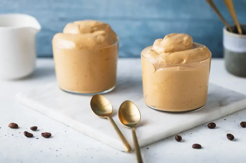 Due bicchieri di soffice crema di caffè, perfetta ricetta crema di caffè senza panna e senza caffè solubile. Due cucchiaini dorati e chicchi di caffè completano la scena.