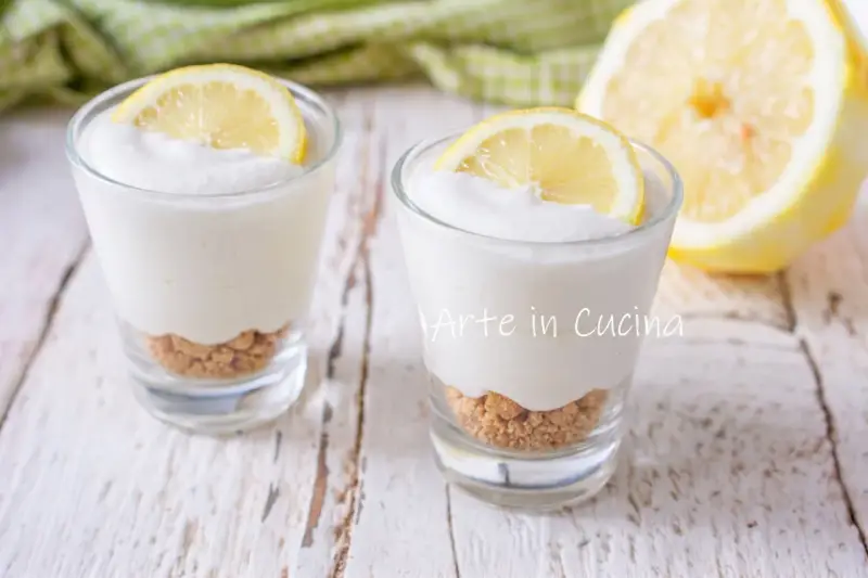 Due bicchierini di mousse al limone con base croccante e fetta di limone, su un tavolo di legno.