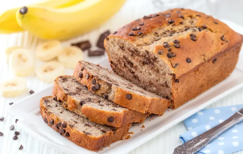 Fette di plumcake banana e cioccolato su un piatto bianco, con banane e gocce di cioccolato sullo sfondo.