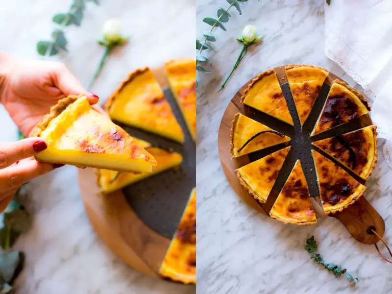 Una mano prende una fetta di flan parisien dorato. La torta intera, tagliata a spicchi, è su un tagliere di legno, decorata con un fiore e un rametto di eucalipto.